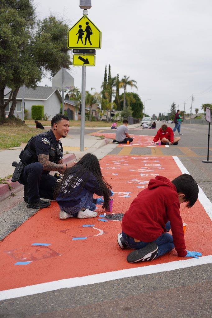 A police officer kneeling to help children paint a crosswalk